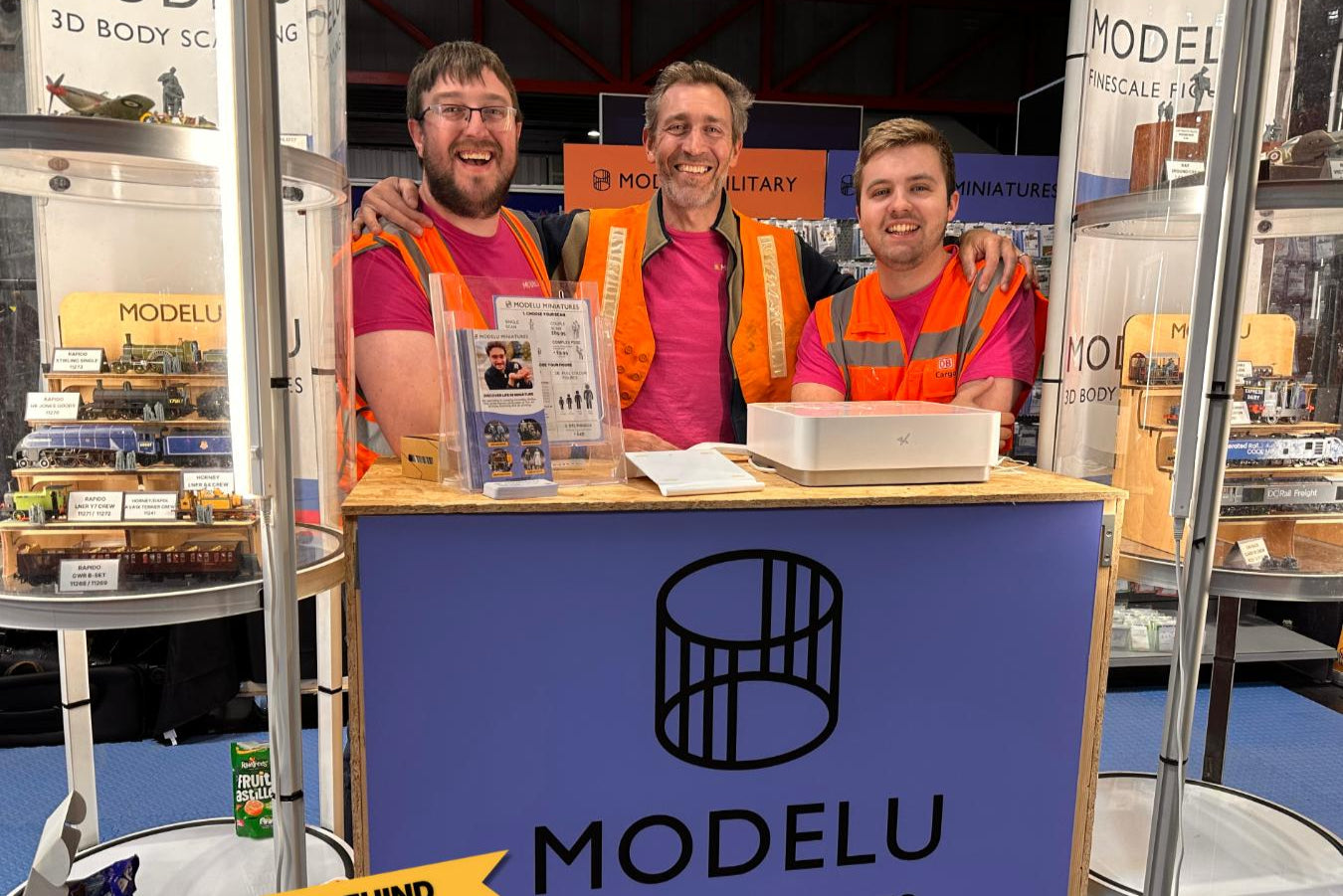 Three men in high-visibility vests behind a Modelu branded booth at an exhibition.