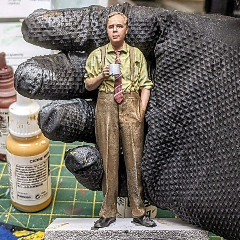 Hand holding a miniature figure on a paint palette with art supplies in the background