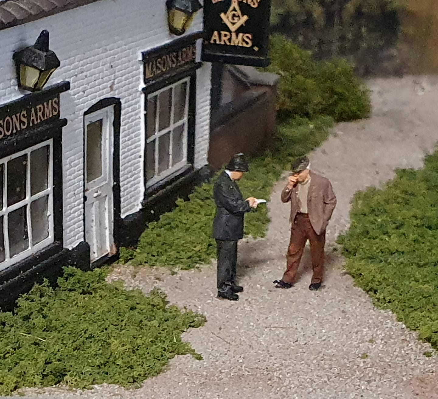 A miniature model scene of a white brick pub named Mason's Arms with two small figurines standing on a gravel path in front of the building.