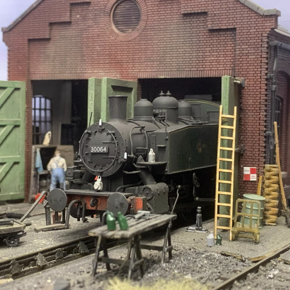 Model train set with a steam locomotive in front of a brick building.