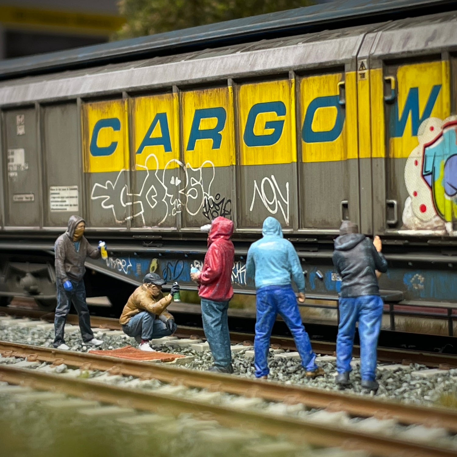 A miniature model freight train car with graffiti and large blue 'CARGO' lettering, set on model railroad tracks with small figurines of people standing beside it.